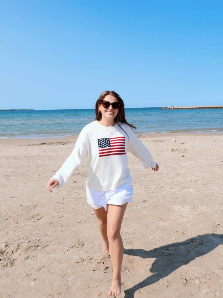 A woman in a white American flag sweatshirt and white shorts walking on a sunny beach.