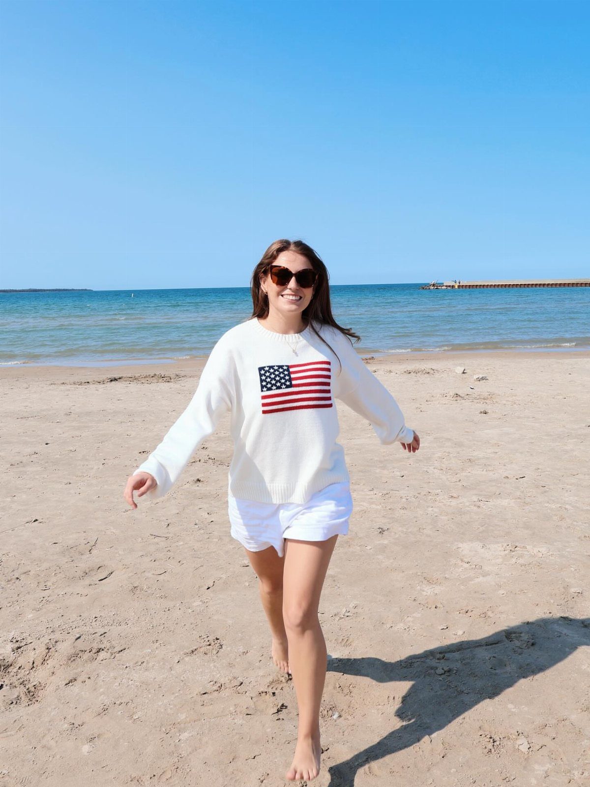 A woman in a white American flag sweatshirt and white shorts walking on a sunny beach.