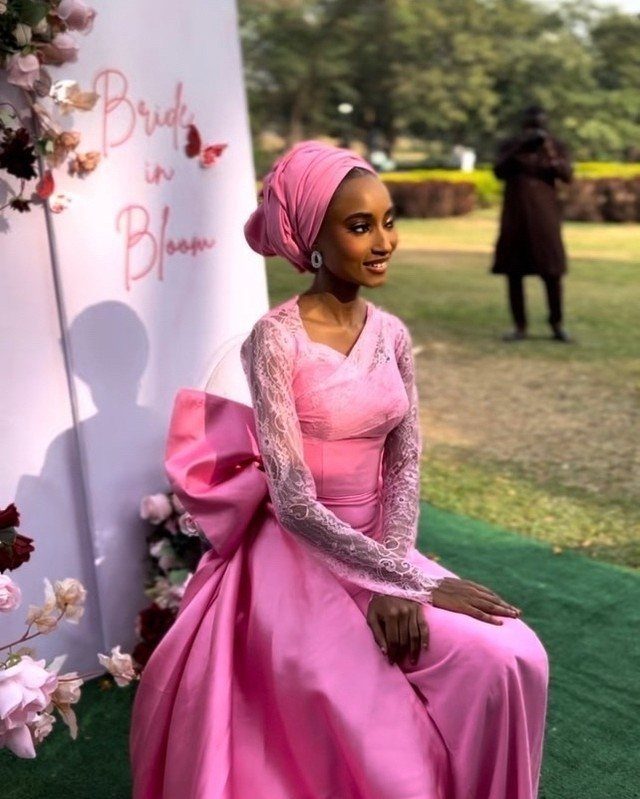 A woman in a bright pink lace gown and matching headwrap sitting outdoors.
