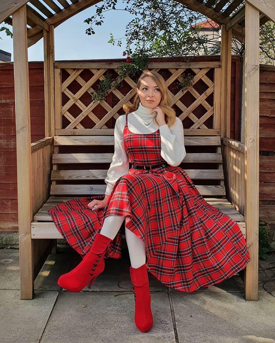 Woman sitting on a wooden bench wearing a red plaid dress and red boots.