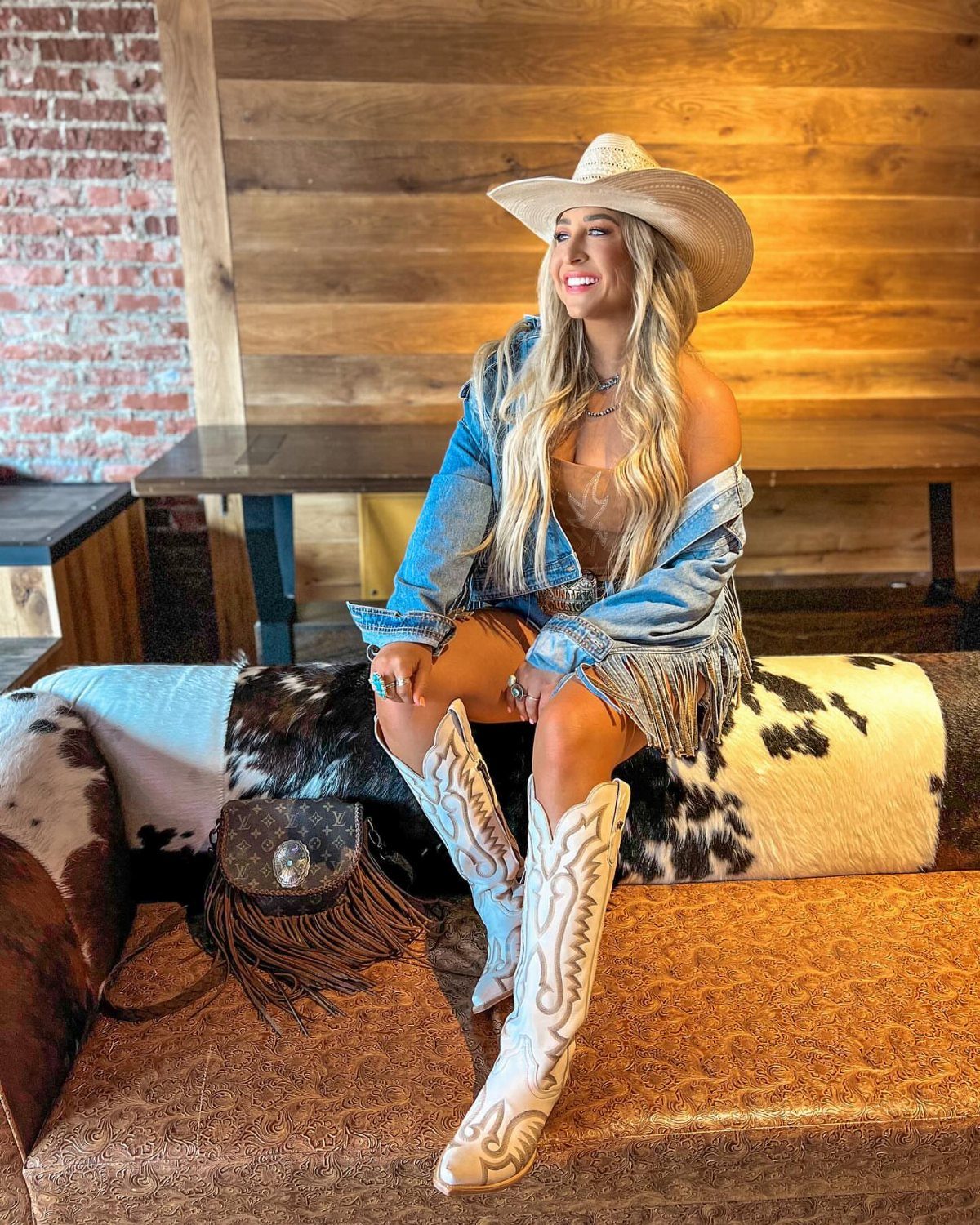 A woman in a denim fringe jacket and white cowboy boots sitting on a cowhide bench.