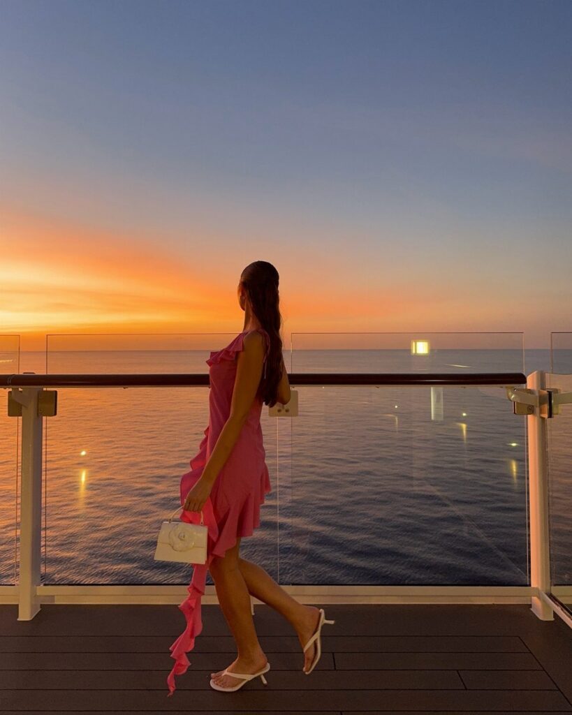 A woman in a pink dress and white sandals stands on a balcony overlooking the ocean sunset.
