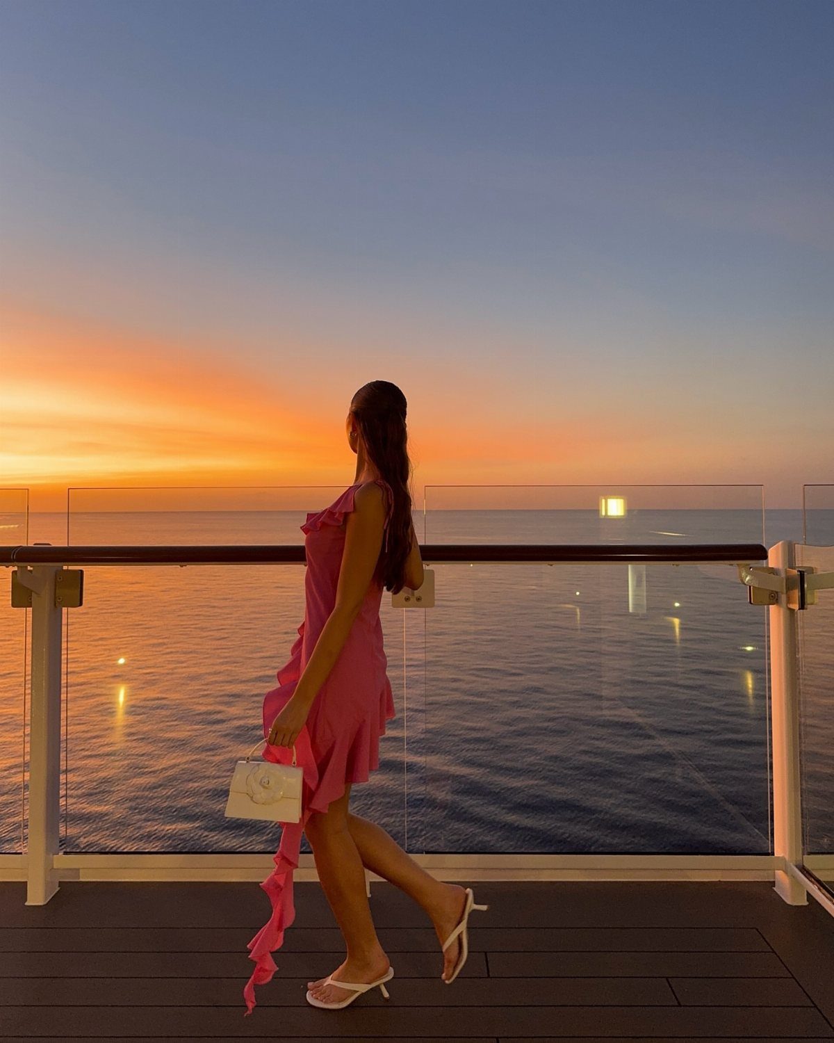 A woman in a pink dress and white sandals stands on a balcony overlooking the ocean sunset.