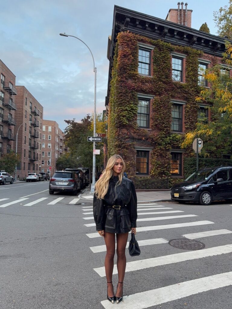 Blonde woman in black leather outfit and heels standing on a city street crosswalk.