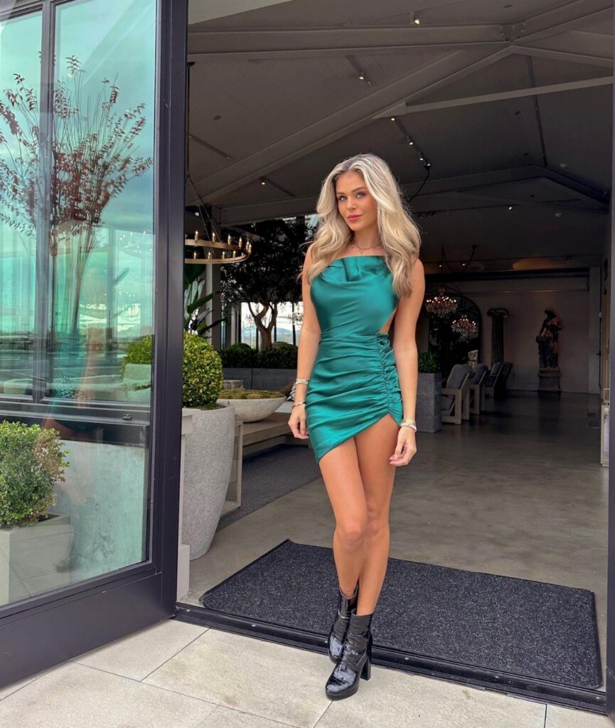 Blonde woman posing in a short emerald green satin dress and black boots.