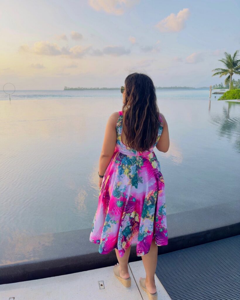 Woman in a colorful floral sundress standing by an infinity pool overlooking the ocean at sunset.