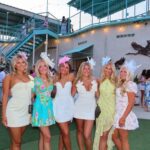 Six women in colorful summer dresses and decorative headpieces posing together outdoors.
