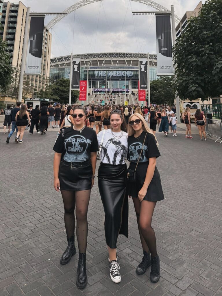 Three women in black graphic tees and leather skirts posing in front of a stadium.