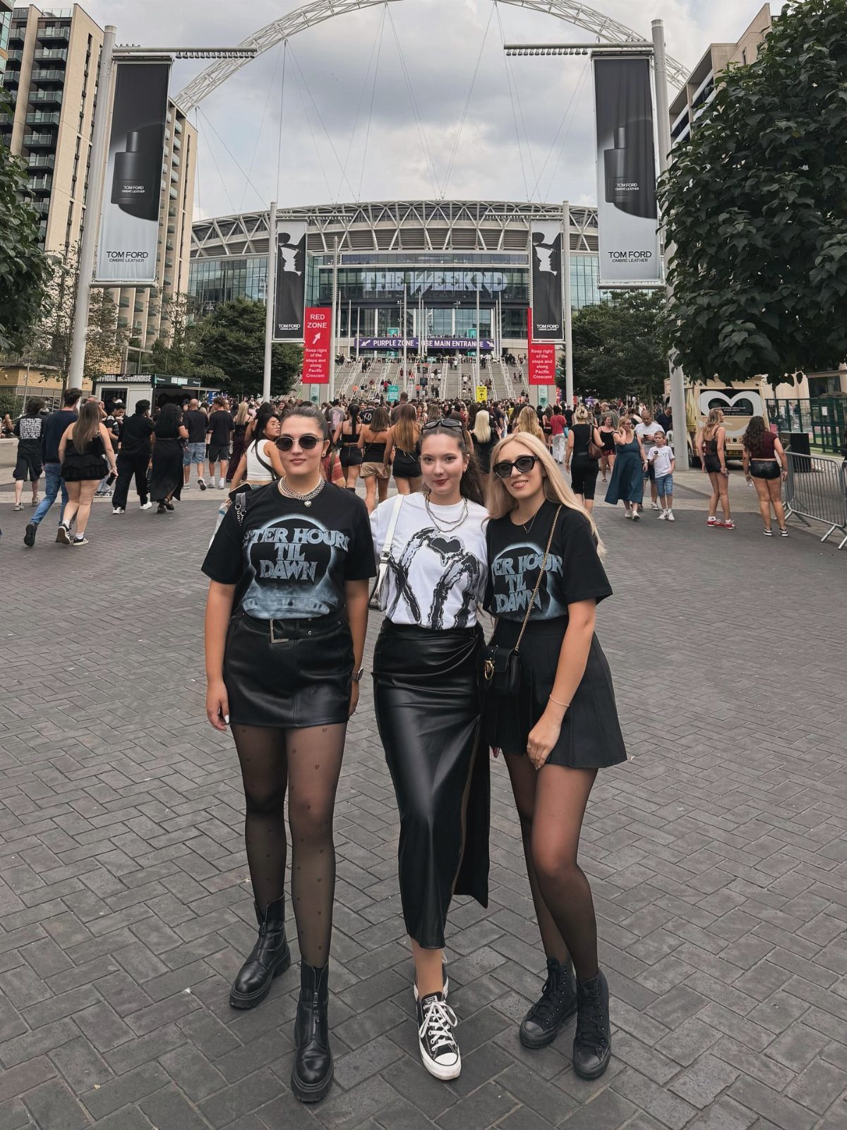 Three women in black graphic tees and leather skirts posing in front of a stadium.