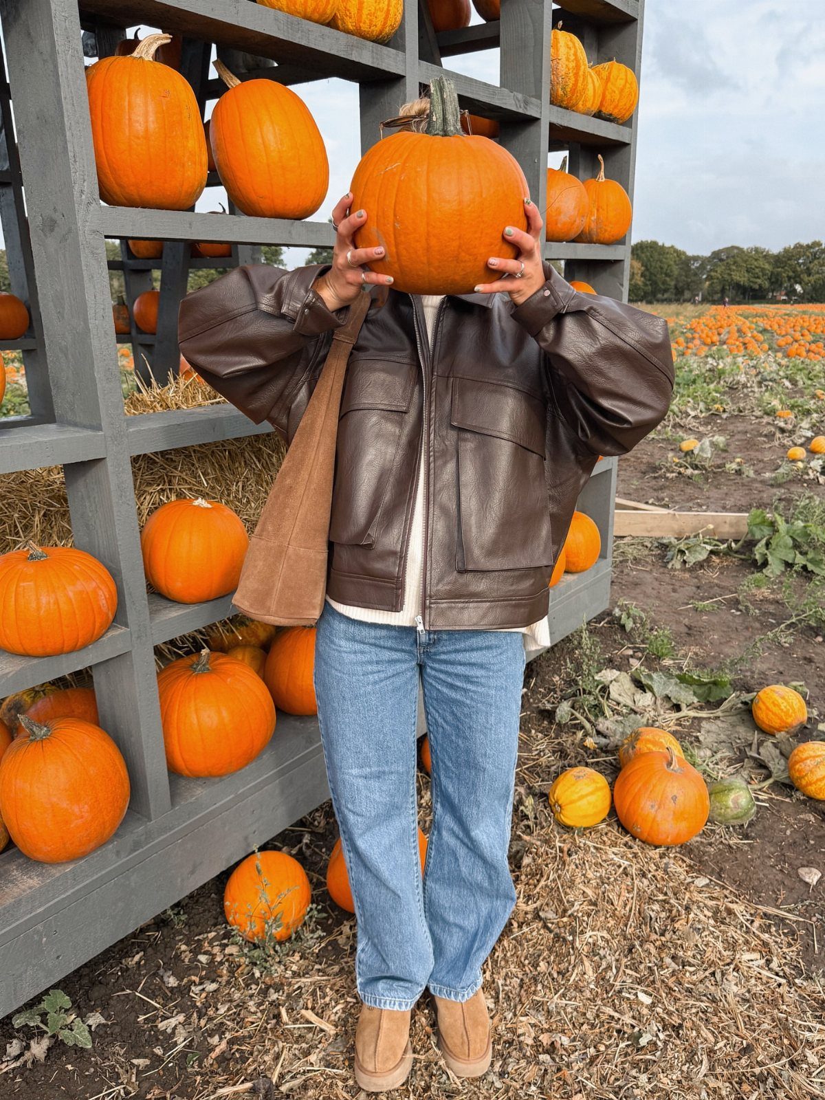 Person in a leather jacket and jeans holding a pumpkin in front of their face.