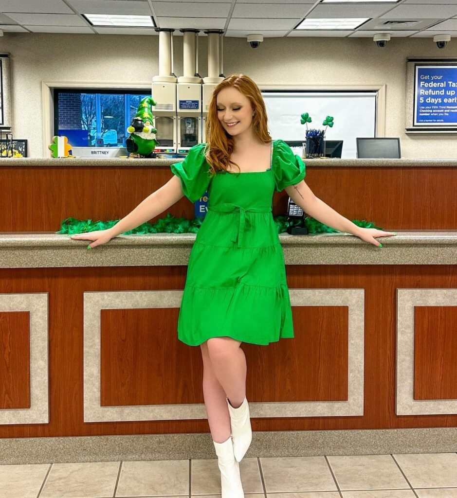 Red-haired woman in a bright green puff-sleeve dress and white boots leaning against a wooden counter.
