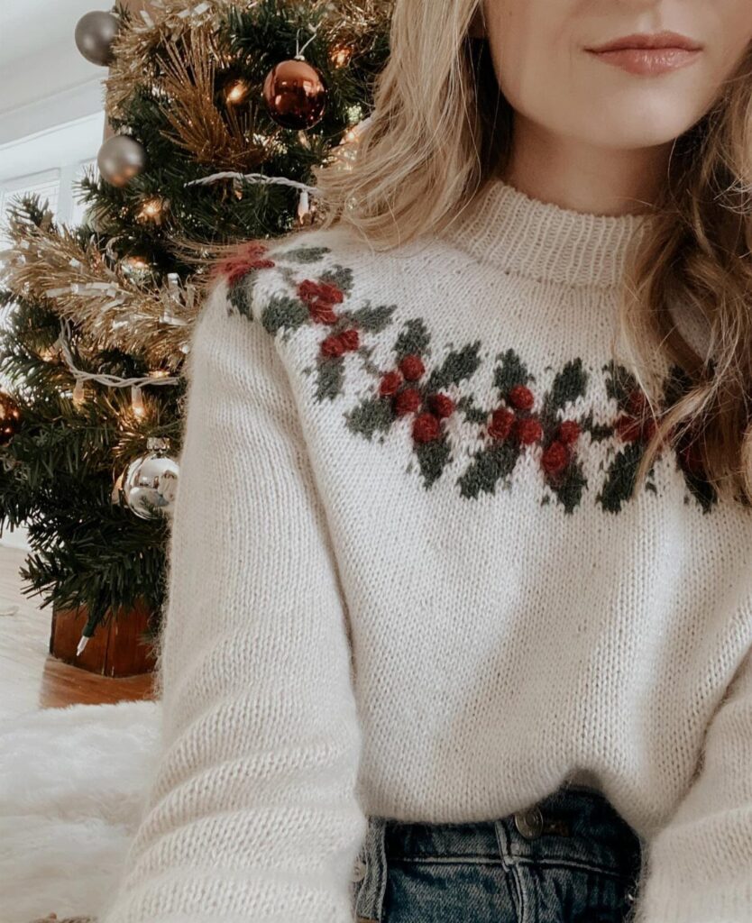 Woman wearing a cream knit sweater with holly berry patterns in front of a Christmas tree.