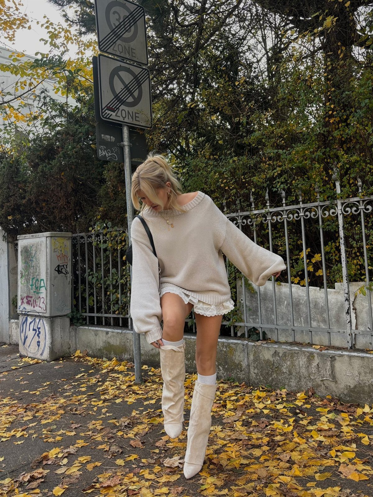 Woman in a cream sweater dress and white knee-high boots standing on autumn leaves.
