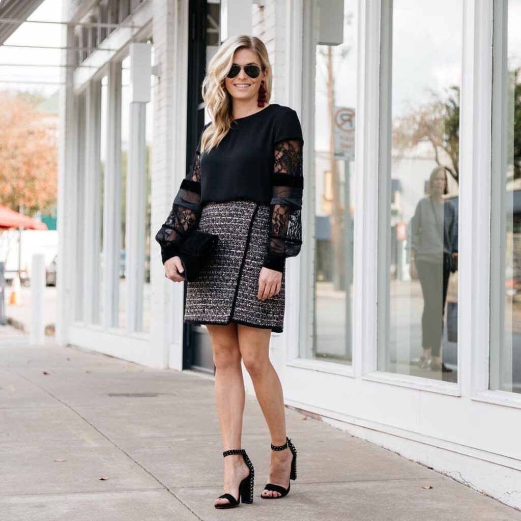 A blonde woman posing in a black lace top, tweed skirt, and black high heels.