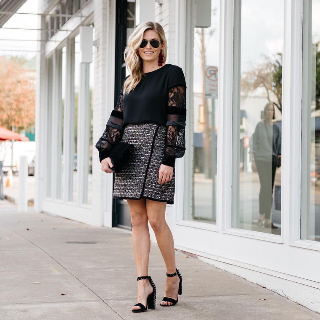 A blonde woman posing in a black lace top, tweed skirt, and black high heels.